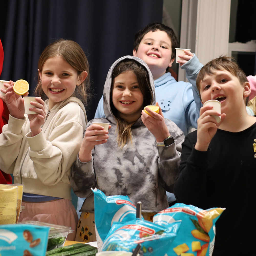 A group of school-aged children holding up fruits at a smoothie workshop.